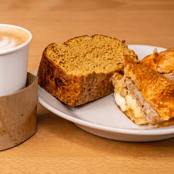 Latte with leaf foam art beside a plate of sliced cake and golden cheese-filled pastries on a wooden table.