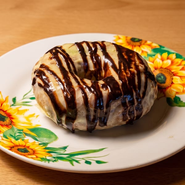 A donut with white frosting and chocolate drizzle on a white plate with sunflower designs.